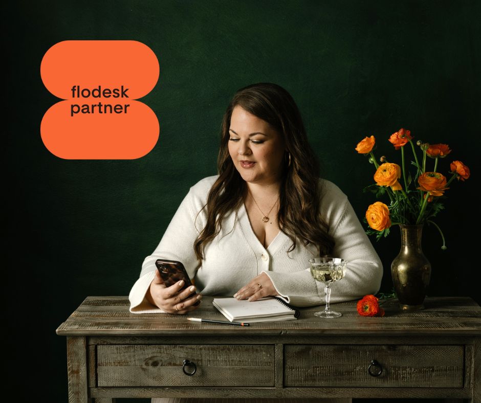 Vanessa Bucceri sitting at a desk with her phone and notebook, beside orange ranunculus flowers, shown with a Flodesk Partner badge for her Flodesk review.