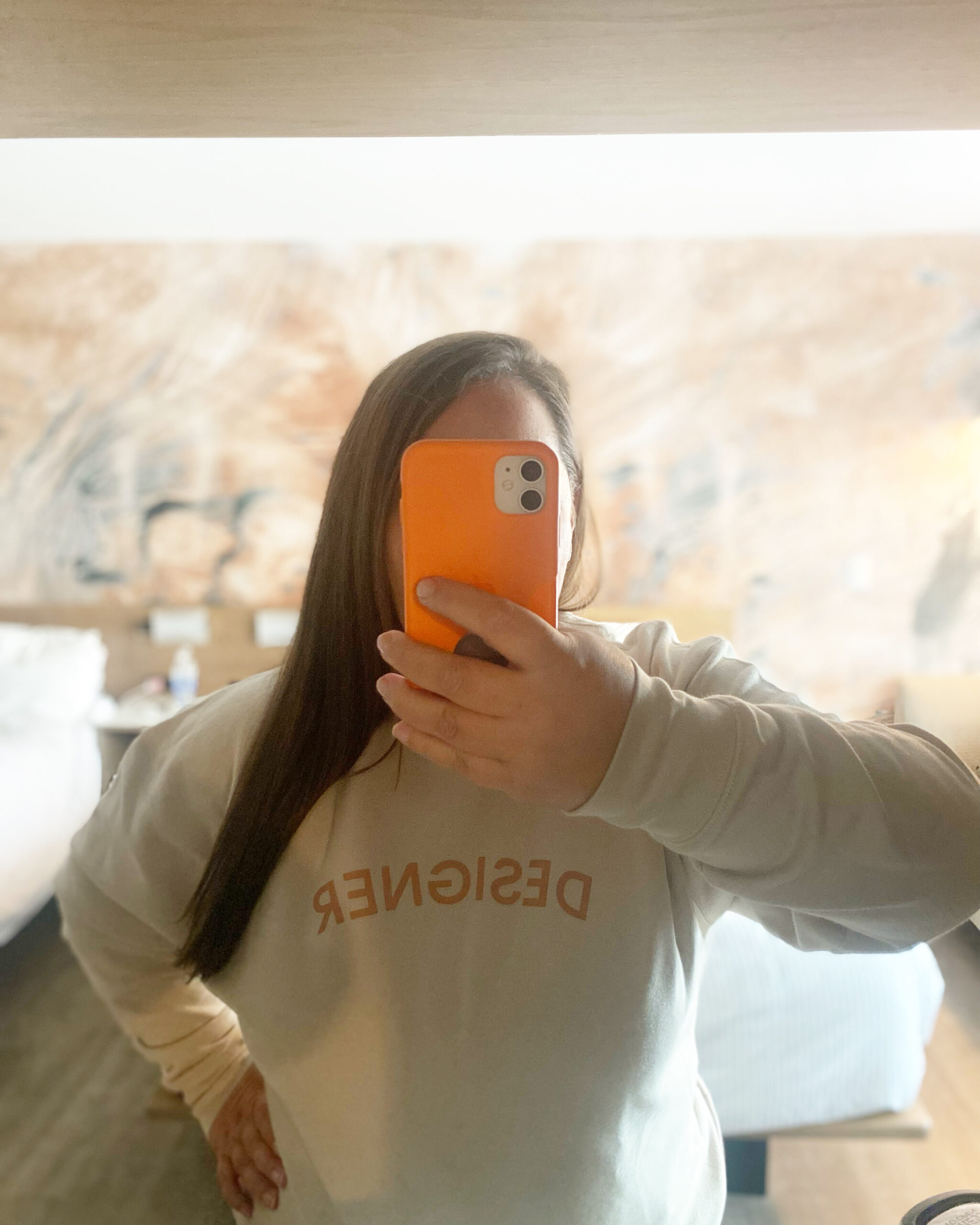 Brand designer Vanessa Bucceri, taking a photo of her reflection in the mirror, her phone covering her face, wearing a sweater that says "Designer"