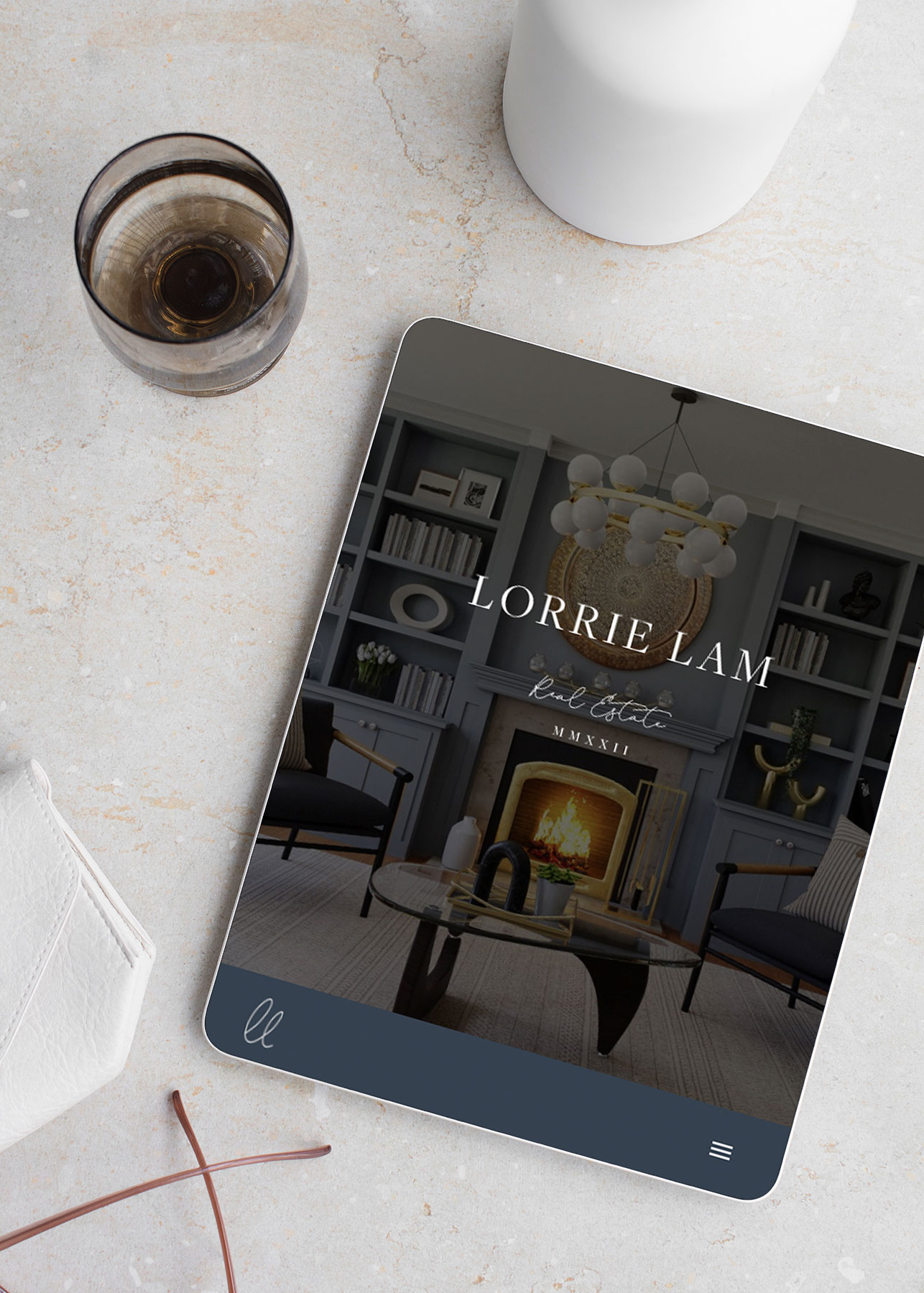 Mockup of Lorrie's real estate website homepage design displayed on a tablet. The design features a luxurious interior with a lit fireplace, built-in shelves, and elegant furniture. The website displays her wordmark logo in a sophisticated serif font with clean, minimalist styling.