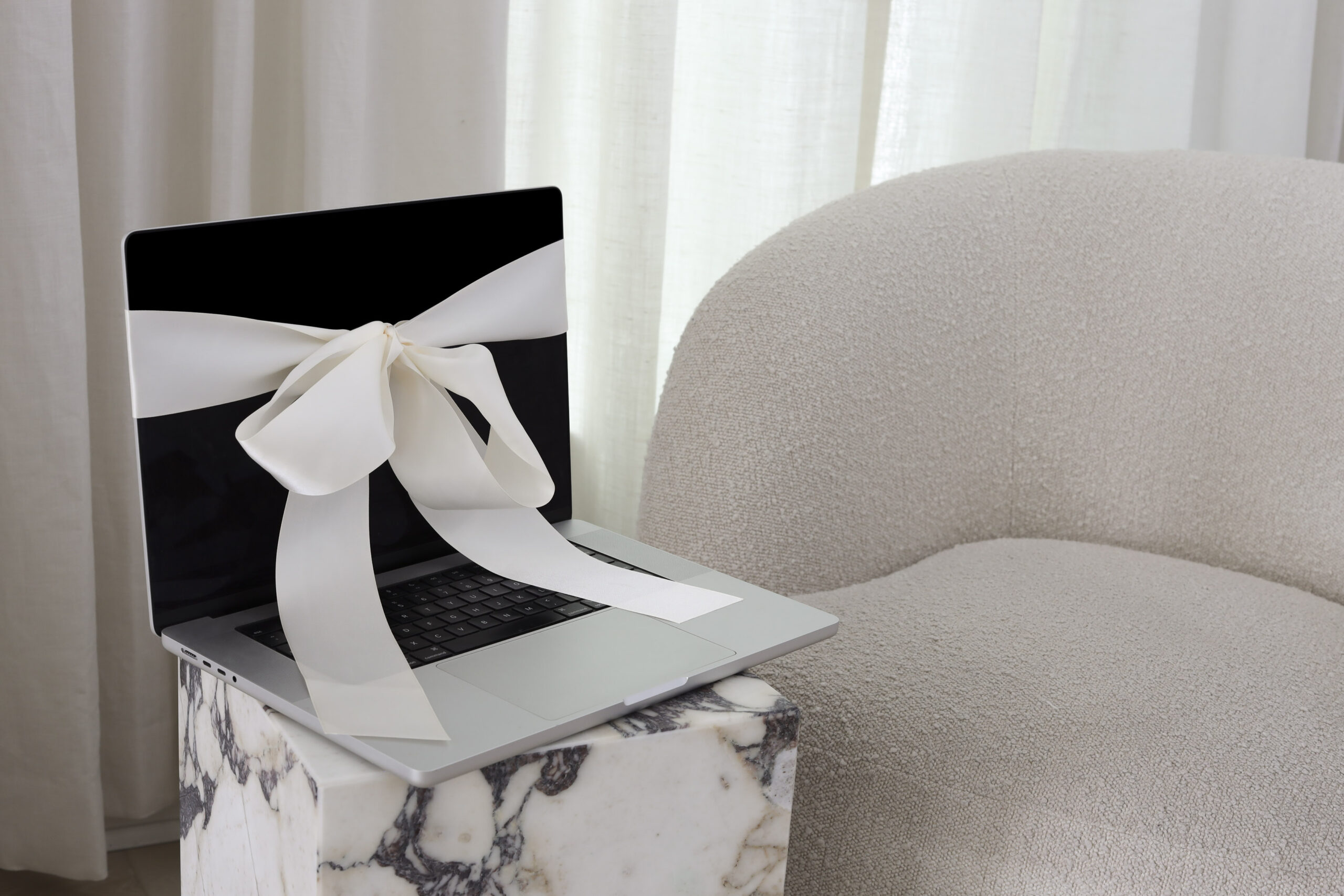 Open laptop wrapped with an elegant white ribbon, sitting on a marble pedestal next to a cozy neutral-toned armchair, symbolizing thoughtful and intentional work in a creative business.